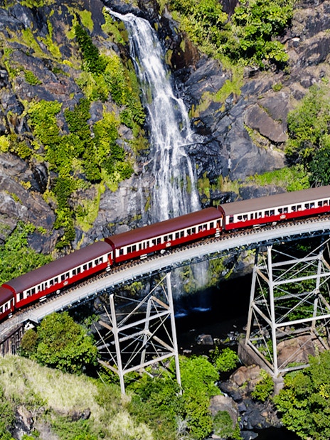 Aerial view of Kuranda Scenic Railway crossing a bridge with a waterfall in the background.