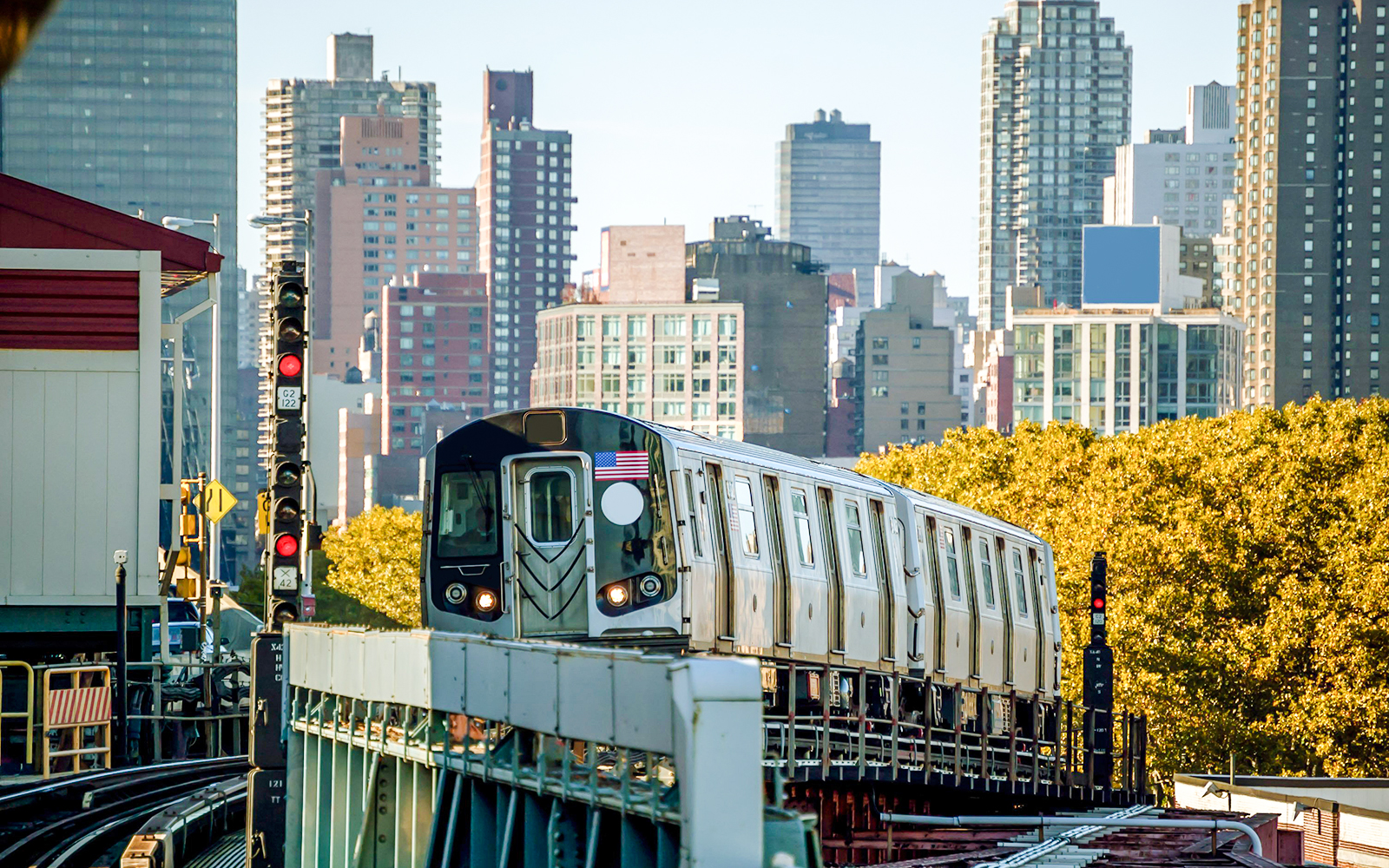 Subway train approaching Bedford Park Boulevard station with city skyline in background.