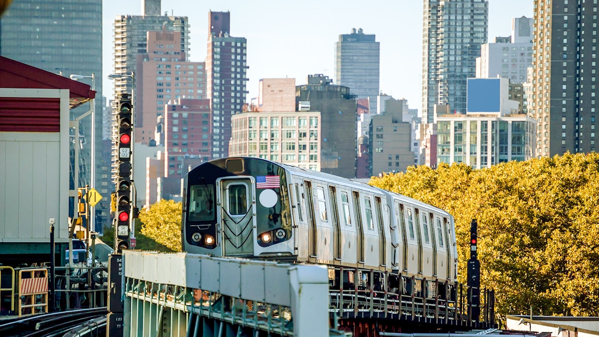 Subway train approaching Bedford Park Boulevard station with city skyline in background.
