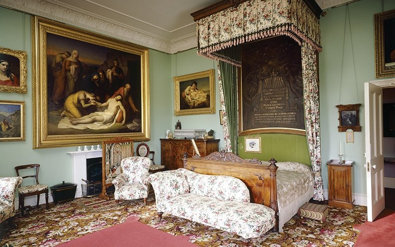 Historic family room with ornate bed, floral upholstery, and classic paintings on green walls.