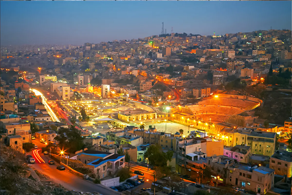 Amman cityscape at dusk with illuminated Roman Theater, viewed from a hilltop.