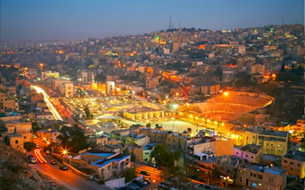 Amman cityscape at dusk with illuminated Roman Theater, viewed from a hilltop.