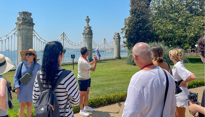 Tour group with guide at Dolmabahce Palace garden, Istanbul, near ornate gate and Bosphorus view.