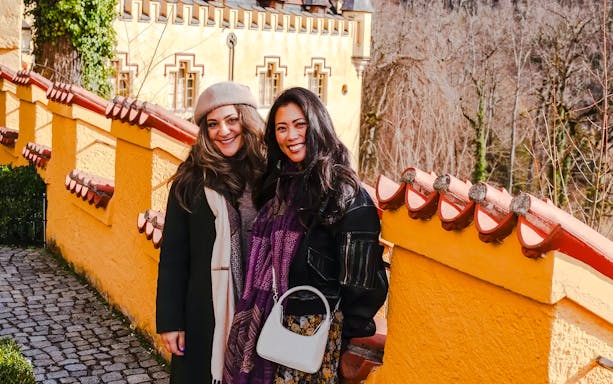 Tourists smiling at Hohenschwangau Castle, Germany, with yellow castle walls in the background.