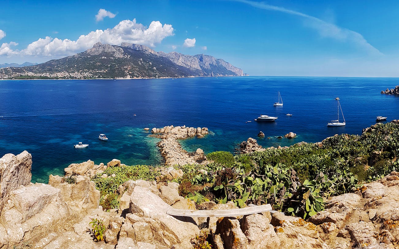 Boats anchored near rocky coastline of Costa di Baunei with mountains in the background.