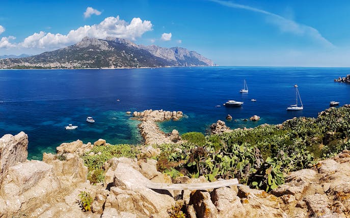 Boats anchored near rocky coastline of Costa di Baunei with mountains in the background.
