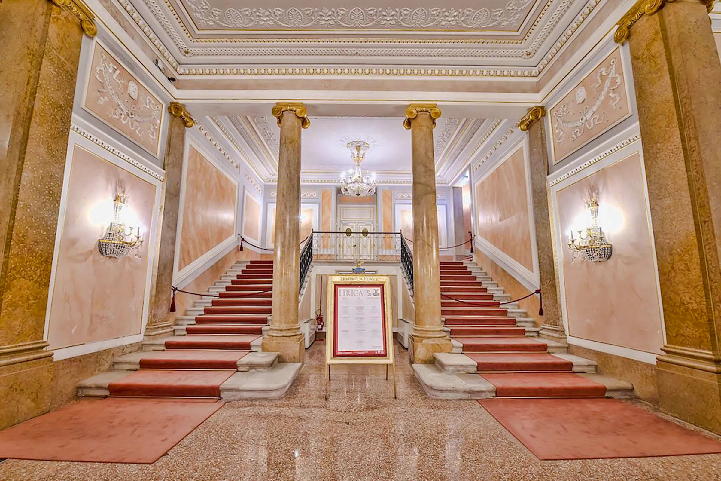 Grand staircase inside La Fenice Opera House, Venice, with ornate decor and chandeliers.
