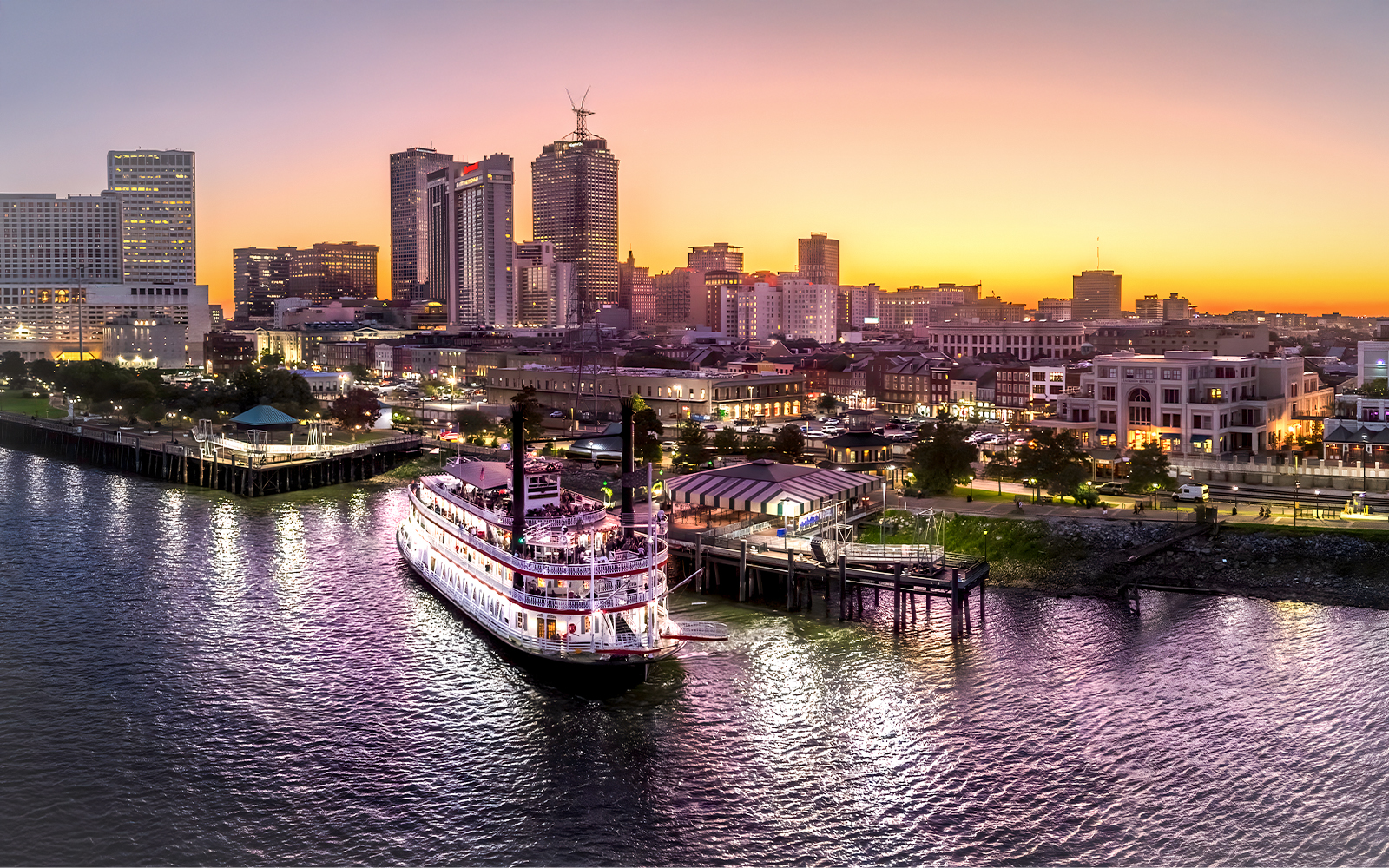 Natchez steamboat on Mississippi River at sunset, New Orleans skyline in background.