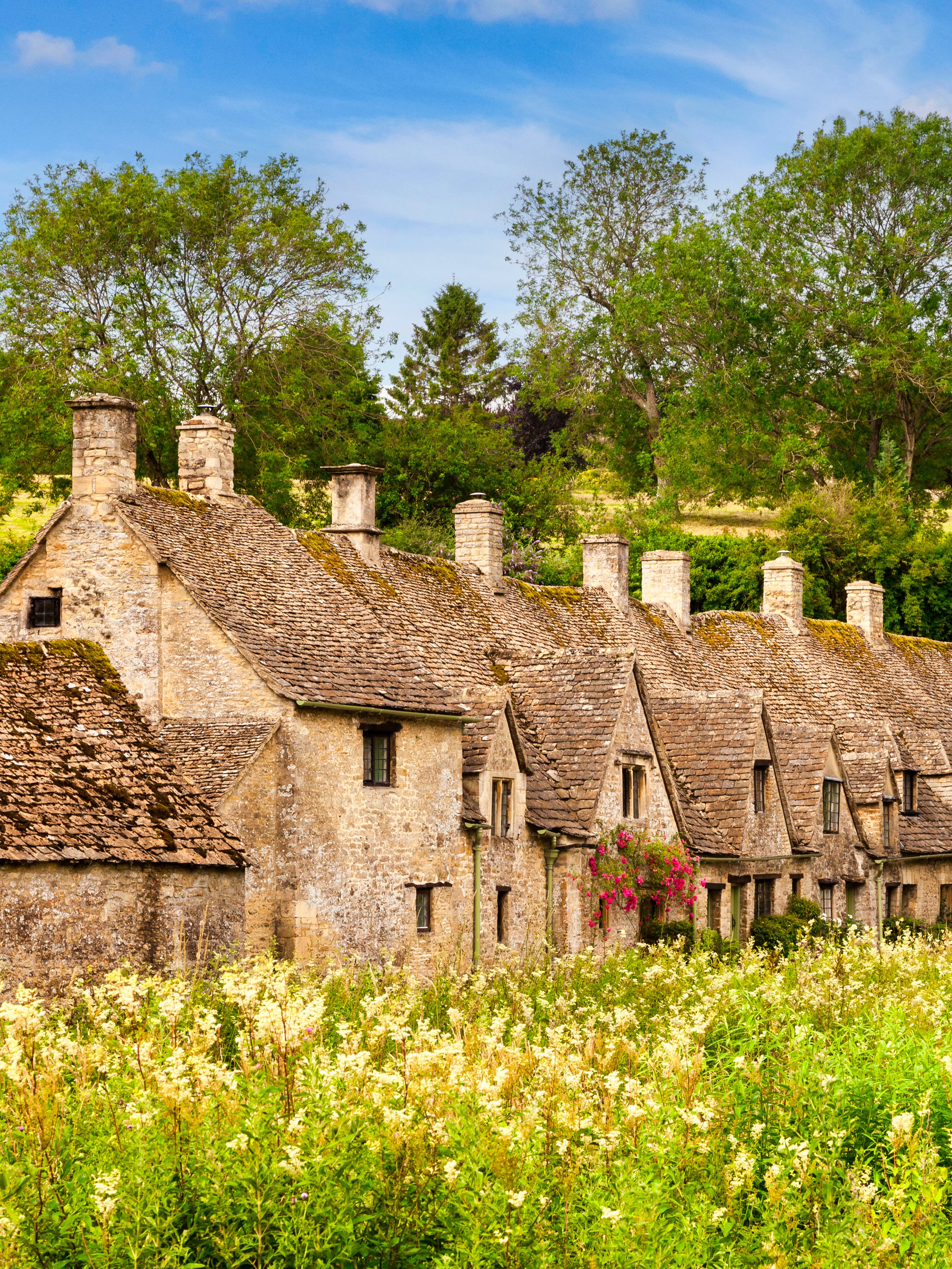 Row of historic stone cottages in Cotswold, England, surrounded by lush greenery.