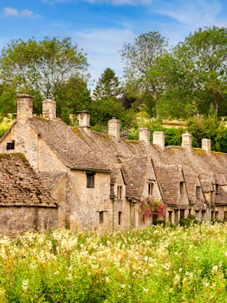Row of historic stone cottages in Cotswold, England, surrounded by lush greenery.