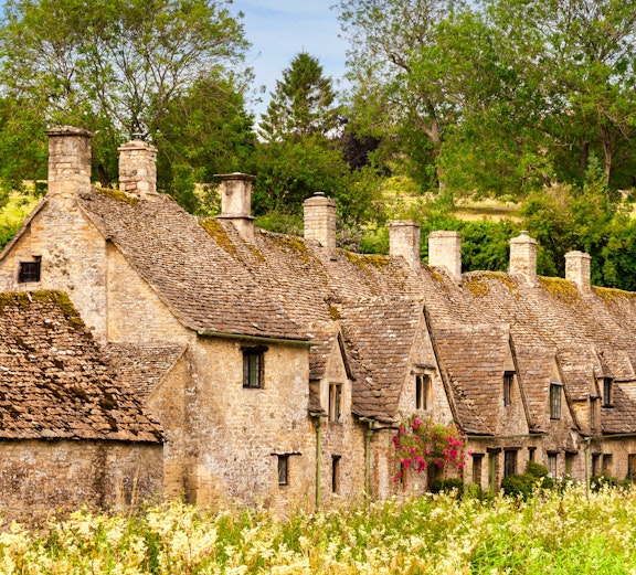 Row of historic stone cottages in Cotswold, England, surrounded by lush greenery.