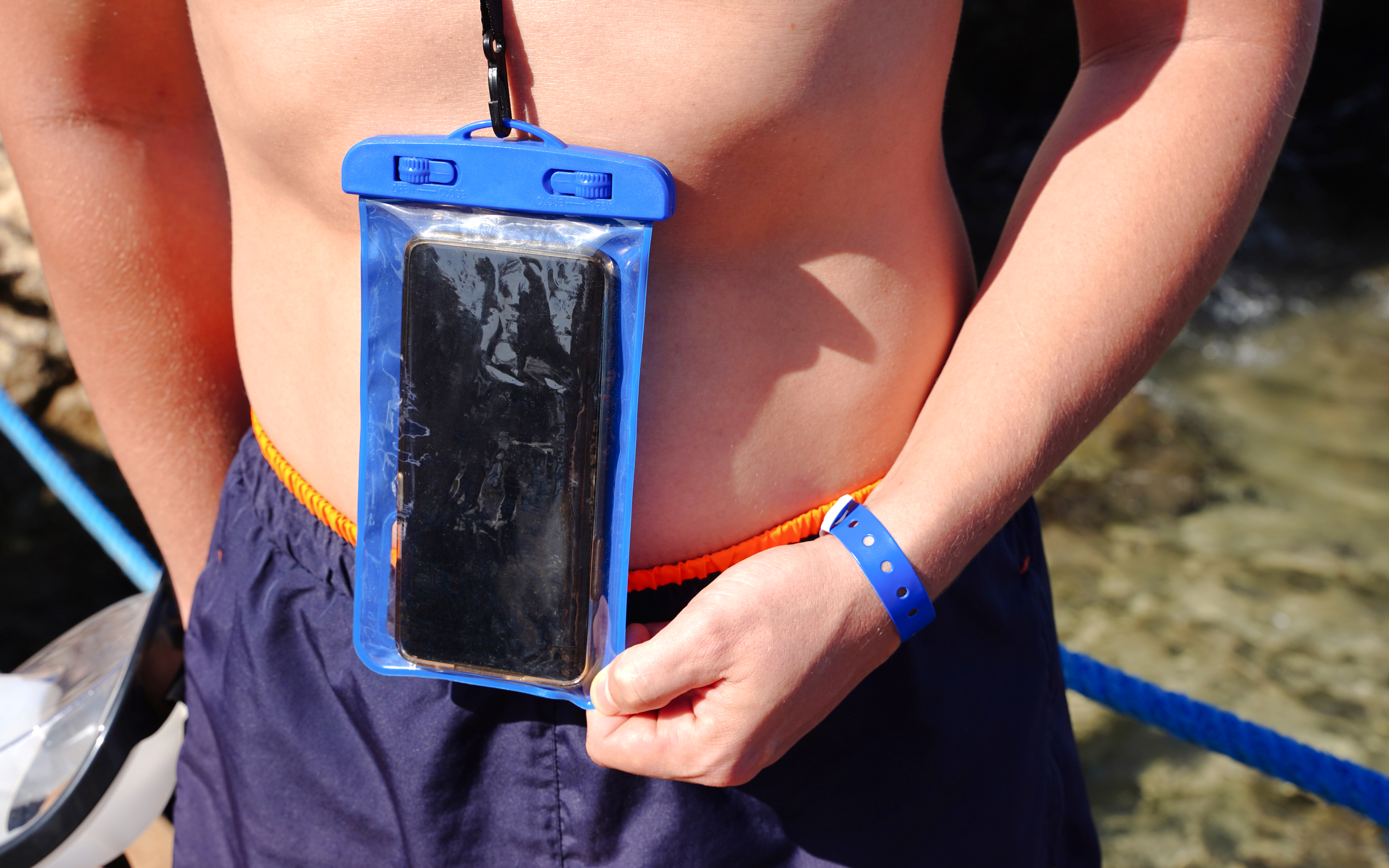 Blue waterproof smartphone case worn around neck near water.