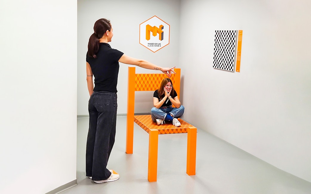 Person pointing at another sitting on oversized chair in perspective room, Museum of Illusions Seville.