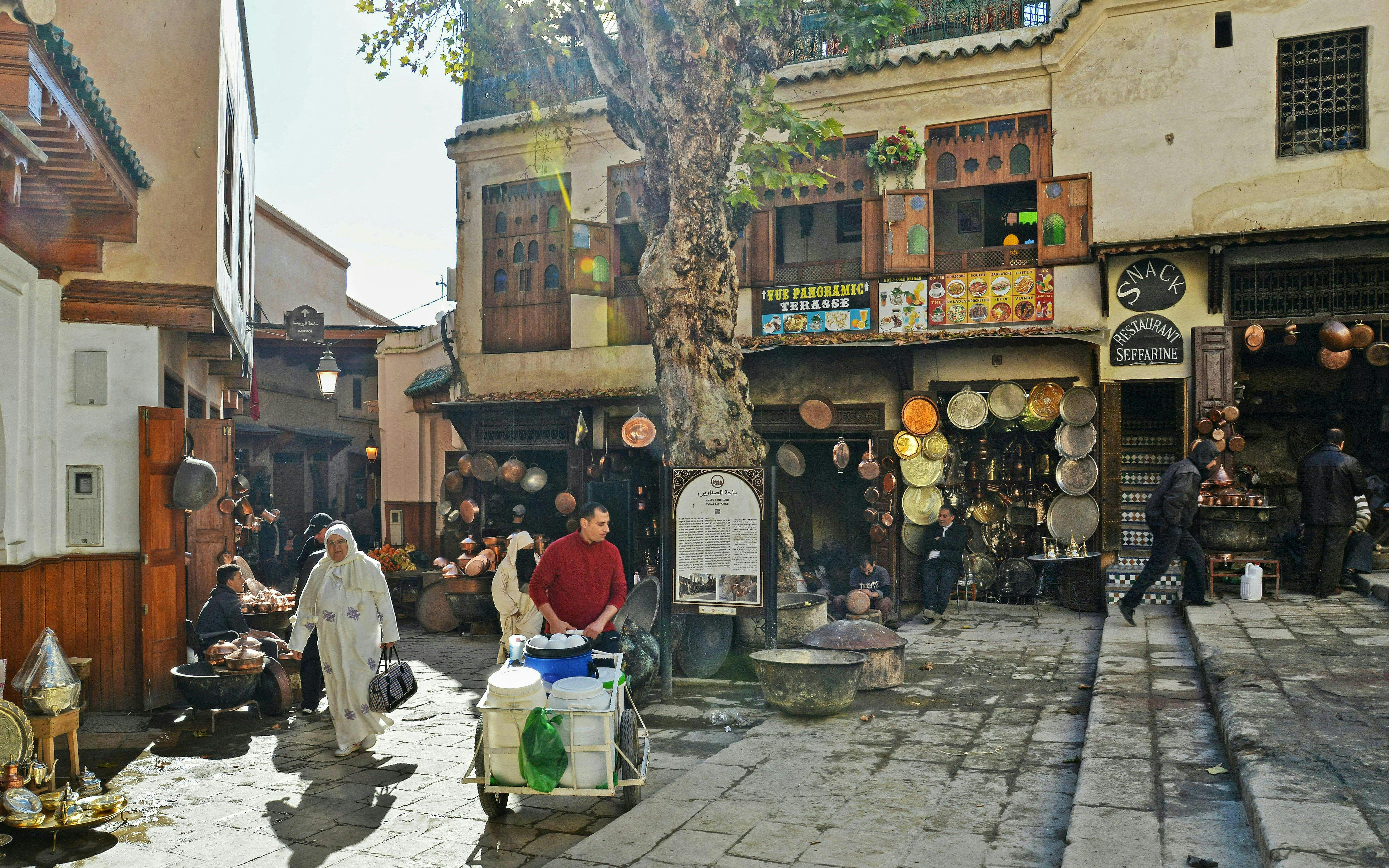 Fes Old Medina market scene with locals, copperware shops, and traditional architecture.