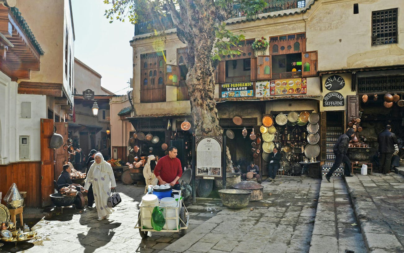 Fes Old Medina market scene with locals, copperware shops, and traditional architecture.