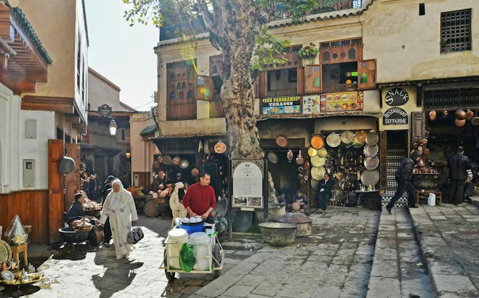 Fes Old Medina market scene with locals, copperware shops, and traditional architecture.