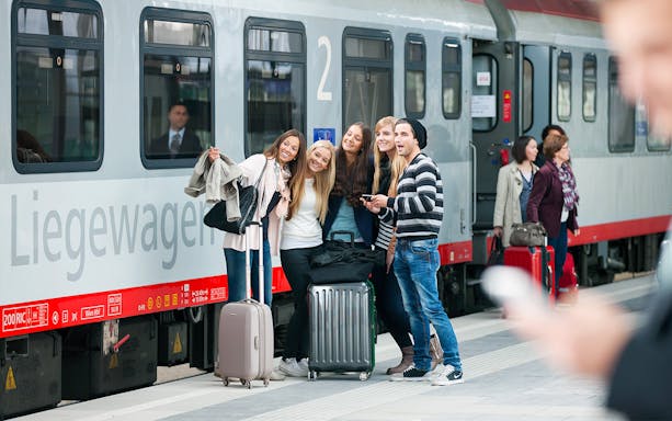 Travellers with luggage at a train station platform next to a Eurail train.