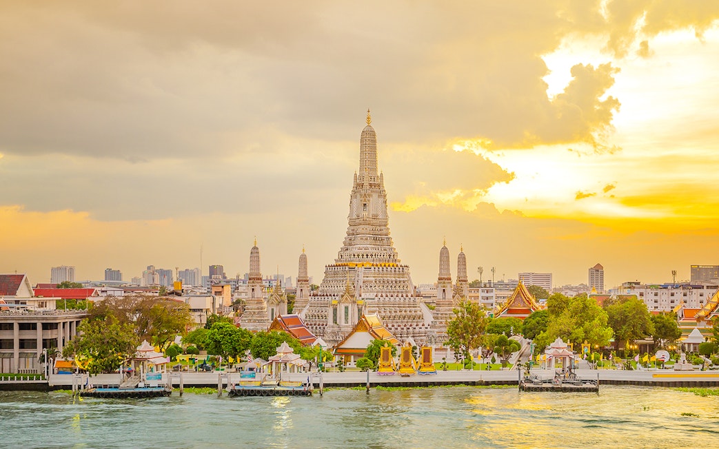 Wat Arun Temple in Bangkok at sunset with Chao Phraya River in the foreground.