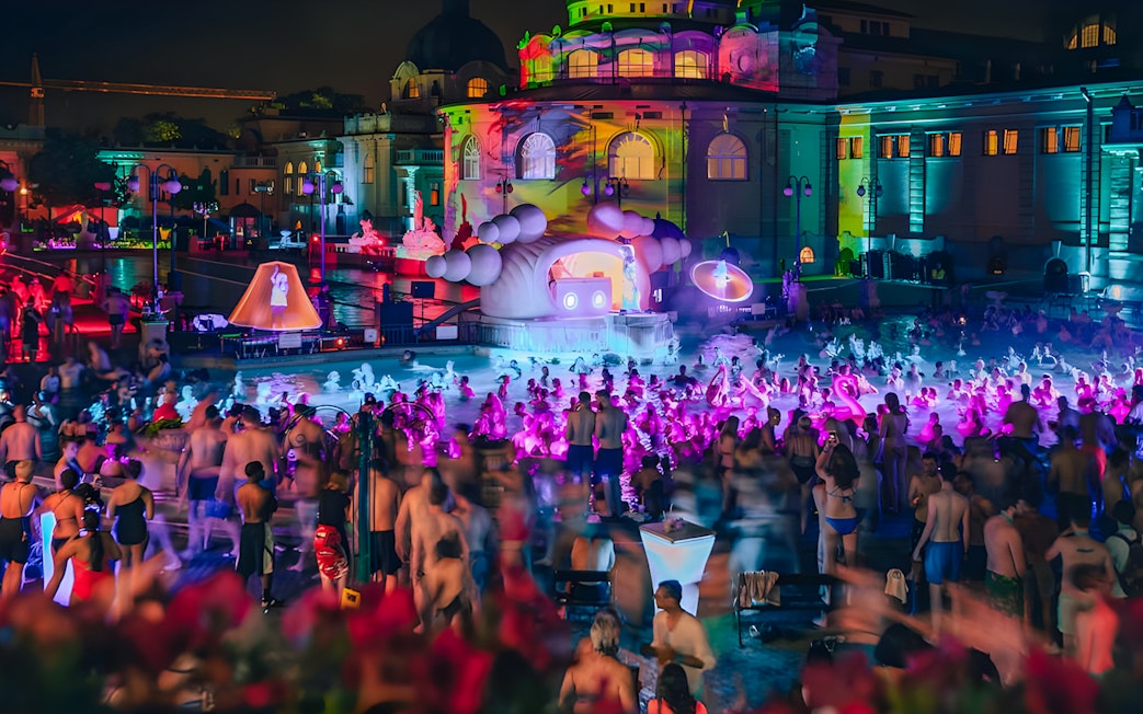 Crowd enjoying a vibrant night pool party with colorful lights at a historic bathhouse.
