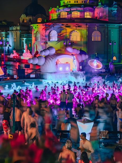Crowd enjoying a vibrant night pool party with colorful lights at a historic bathhouse.