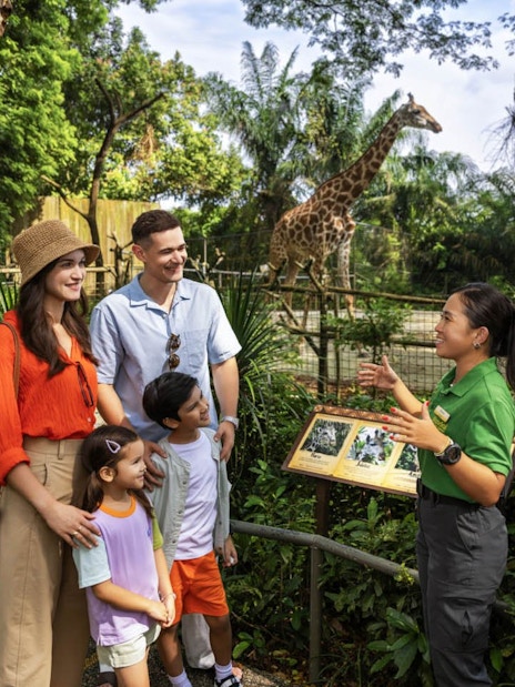 Family with guide at Mandai Wildlife Reserve, giraffe in background.