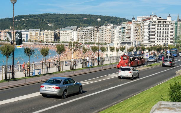 Red tourist train along San Sebastian beach promenade with cityscape in the background.