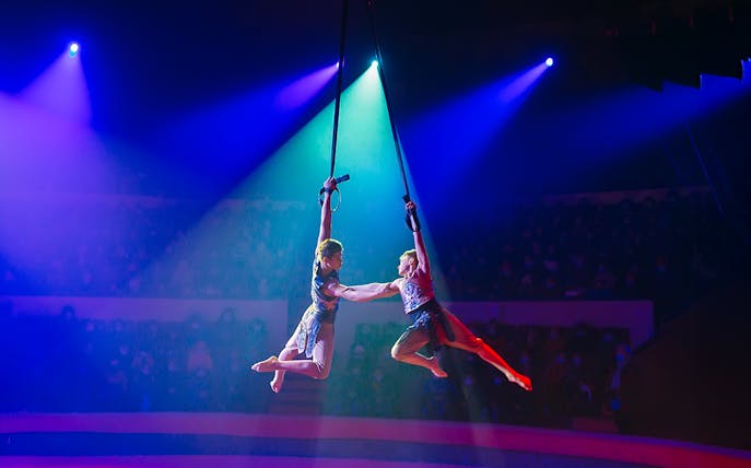 Two boys perform aerial acrobatics on stage under colorful lights.