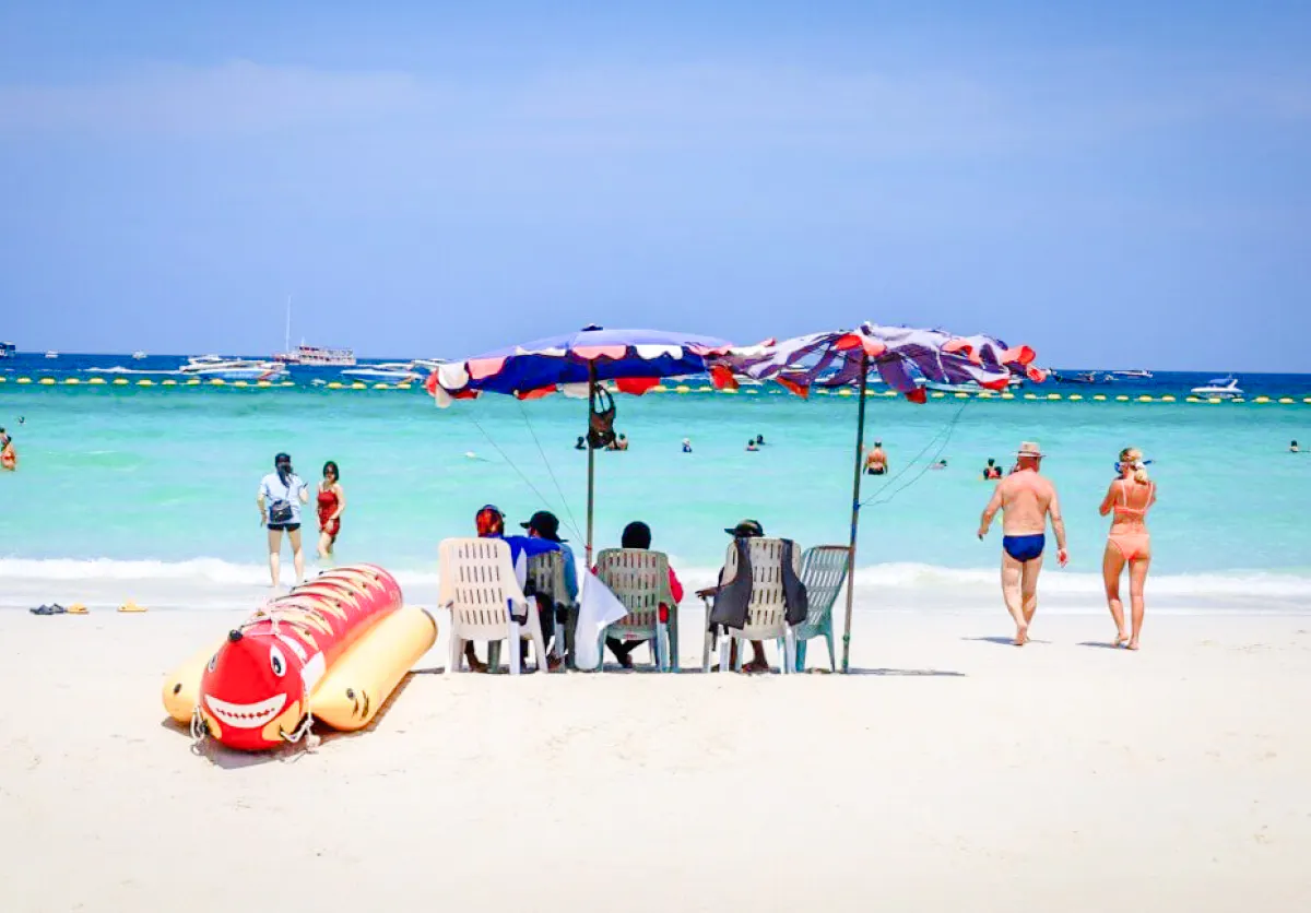 Beachgoers relax under umbrellas on Coral Island beach, Pattaya, with speedboats in the distance.