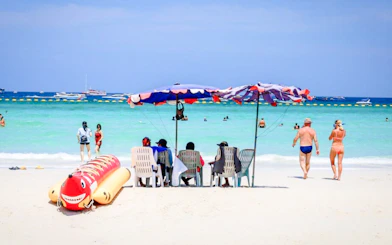 Beachgoers relax under umbrellas on Coral Island beach, Pattaya, with speedboats in the distance.