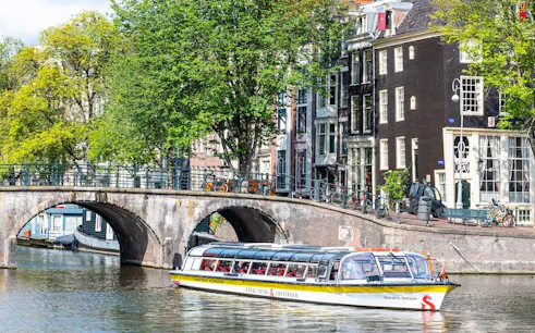 Amsterdam canal cruise boat passing under a stone bridge with historic buildings in the background.