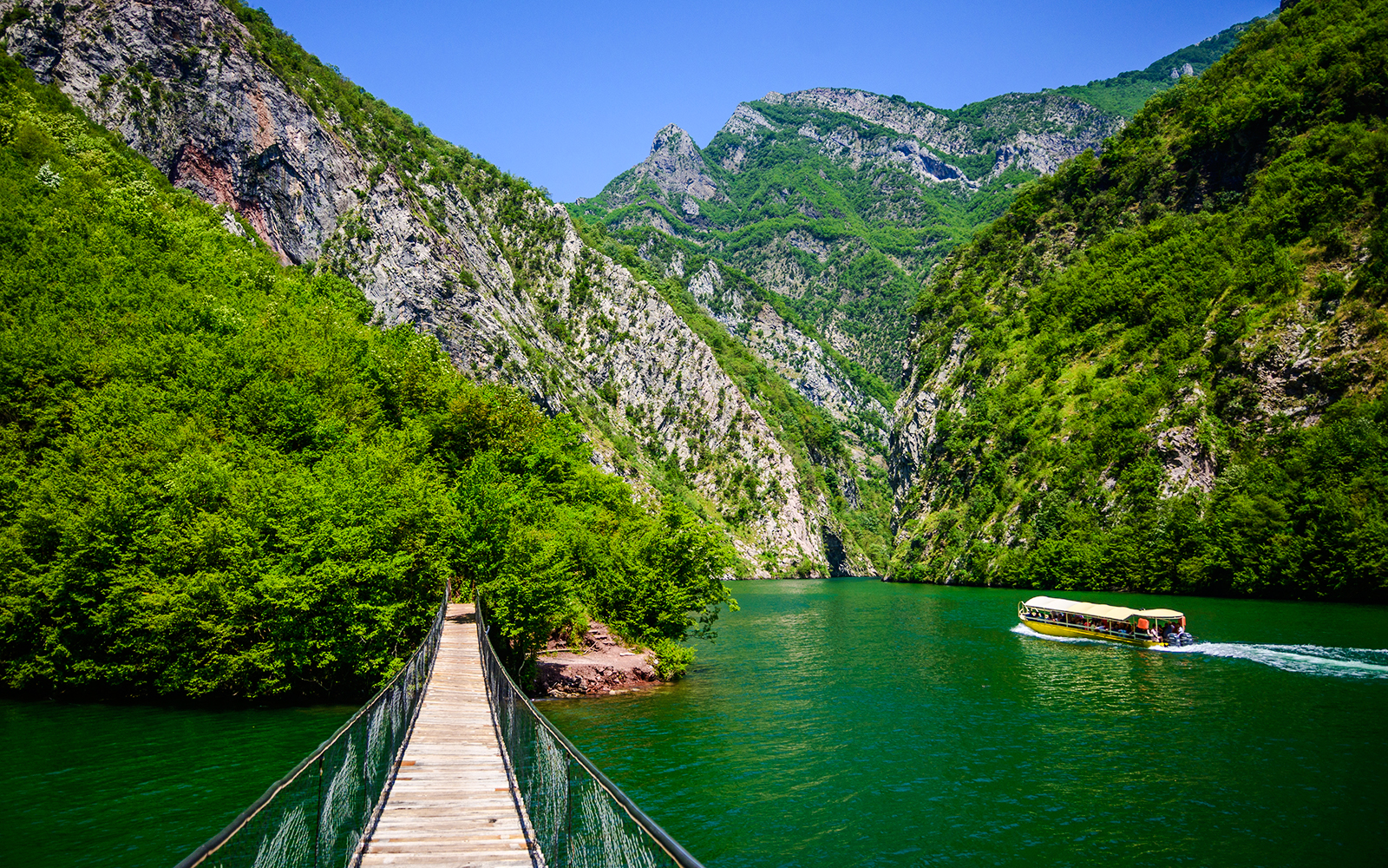 Wooden bridge over Shala River with boat and lush mountains in Tirana.