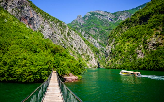 Wooden bridge over Shala River with boat and lush mountains in Tirana.
