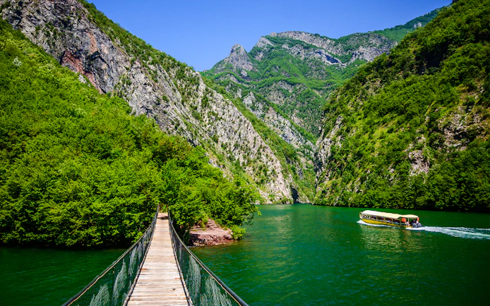 Wooden bridge over Shala River with boat and lush mountains in Tirana.