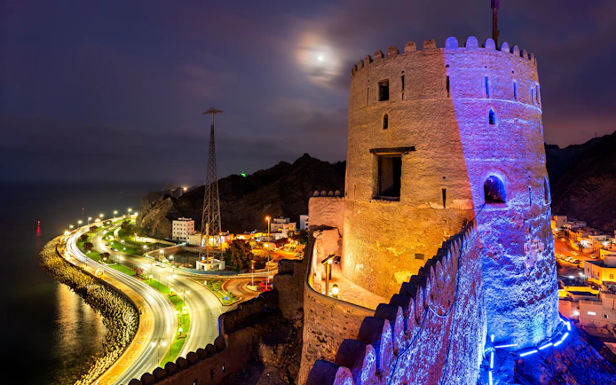 Corniche road and illuminated fort at night in Muscat, Oman.