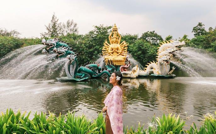 Woman in traditional attire at Erawan Museum with dragon sculptures and golden deity statue.