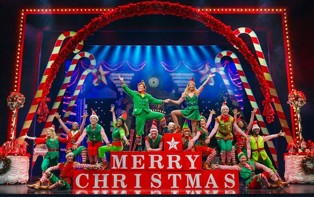 Performers in festive costumes on stage during Elf The Musical, with "Merry Christmas" blocks.