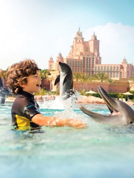 Child interacting with dolphins at Atlantis Waterpark, Dubai, with resort in background.