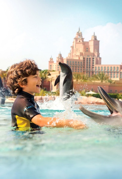 Child interacting with dolphins at Atlantis Waterpark, Dubai, with resort in background.