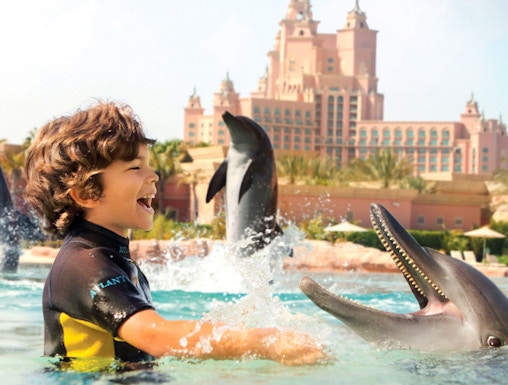 Child interacting with dolphins at Atlantis Waterpark, Dubai, with resort in background.
