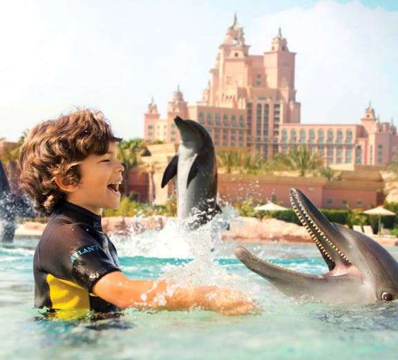Child interacting with dolphins at Atlantis Waterpark, Dubai, with resort in background.