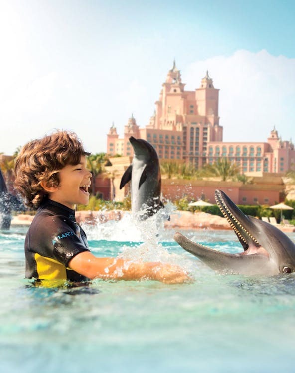 Child interacting with dolphins at Atlantis Waterpark, Dubai, with resort in background.