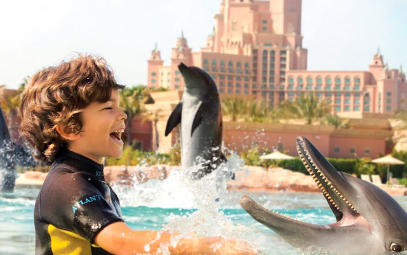 Child interacting with dolphins at Atlantis Waterpark, Dubai, with resort in background.
