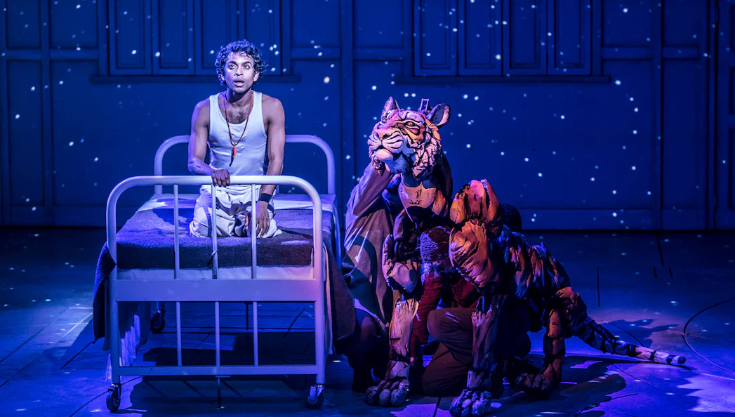 Stage scene from Life of Pi with actor on bed and tiger puppet under starry backdrop.