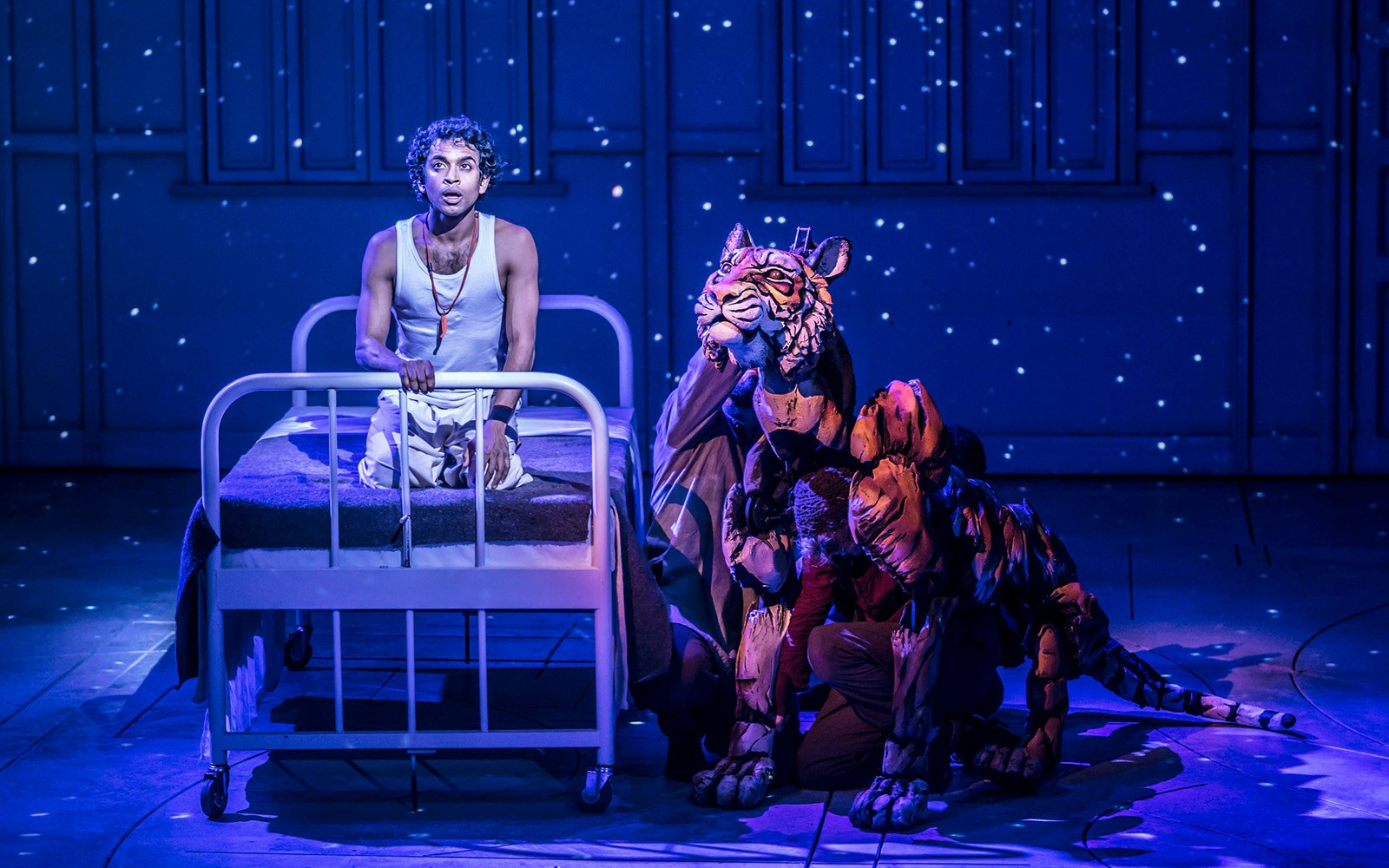 Stage scene from Life of Pi with actor on bed and tiger puppet under starry backdrop.