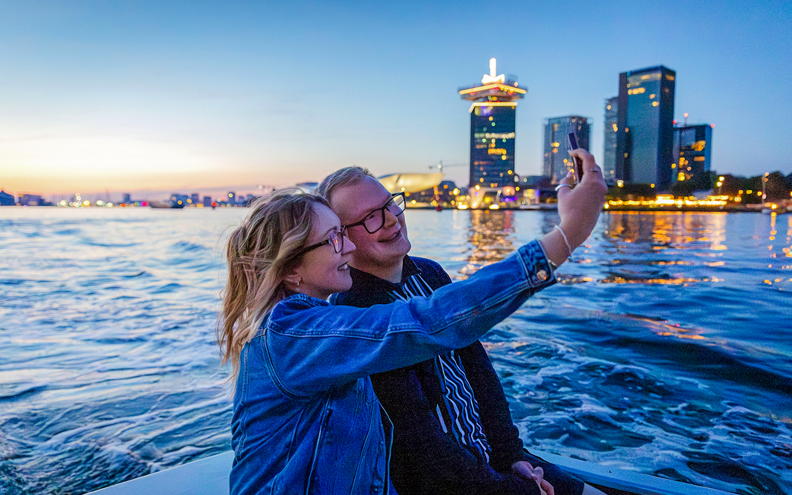 Guests taking a selfie on a winter evening cruise in Amsterdam with city skyline in the background.