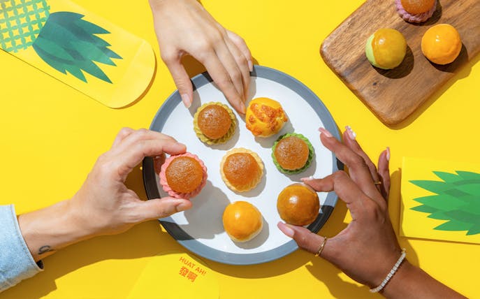 Hands reaching for KELE Singapore pineapple tarts on a plate, with yellow packaging nearby.