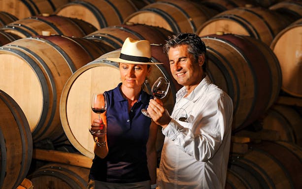 Couple tasting wine in a cellar with wooden barrels in the background.
