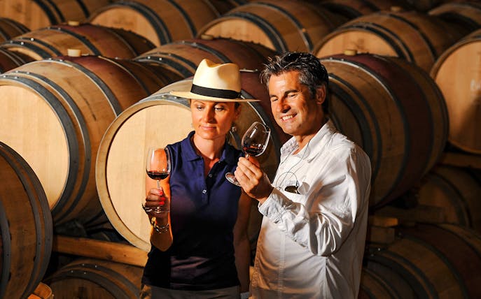 Couple tasting wine in a cellar with wooden barrels in the background.