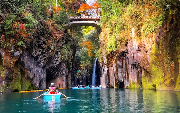 Rowboats navigating Takachiho Gorge with a waterfall and stone bridge in the background, Japan.