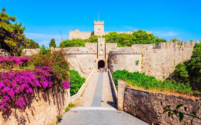 Palace of the Grand Master entrance with stone walls and vibrant flowers, Rhodes, Greece.